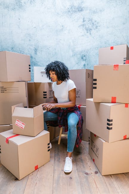 A woman with curly dark hair, dressed in a white t-shirt, blue jeans, and white sneakers, is sitting on a wooden chair surrounded by numerous cardboard boxes during a home relocation or packing proces