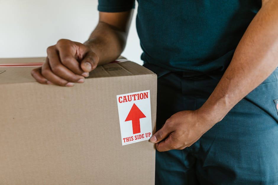 A woman with curly dark hair, dressed in a white t-shirt, blue jeans, and white sneakers, is sitting on a wooden chair surrounded by numerous cardboard boxes during a home relocation or packing proces