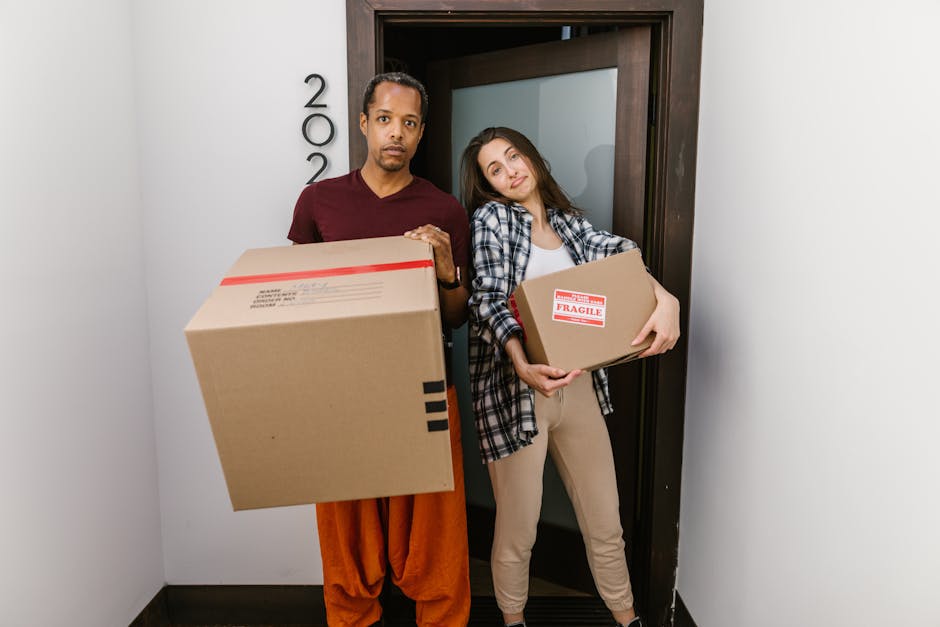 A man and a woman standing inside a property near a door, each holding cardboard boxes used for home relocation. The man is wearing an orange pair of trousers and a maroon shirt, carrying a large card