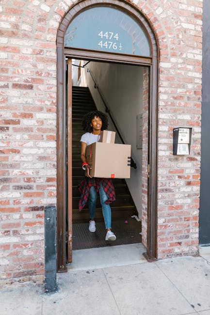 A man and a woman standing inside a property near a door, each holding cardboard boxes used for home relocation. The man is wearing an orange pair of trousers and a maroon shirt, carrying a large card