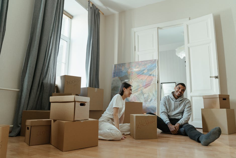 Inside a room prepared for house removals, several cardboard moving boxes are stacked against a wooden wall, with one box secured using clear packing tape. To the left, a white sofa covered with a pro